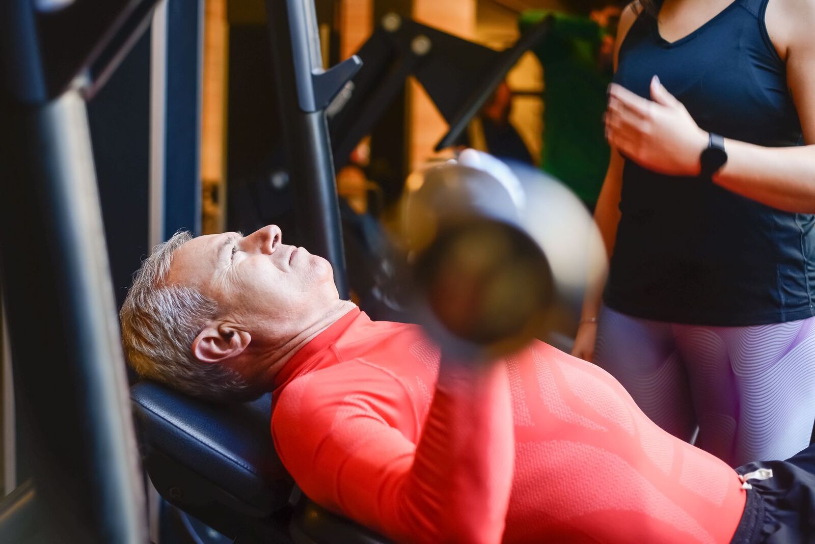 An older man performs a bench press while a coach supervises beside him.